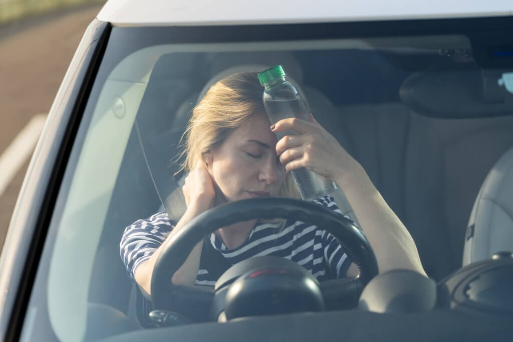 Person sitting at the steering wheel in a car with a bottle of water held to forehead feeling hot.