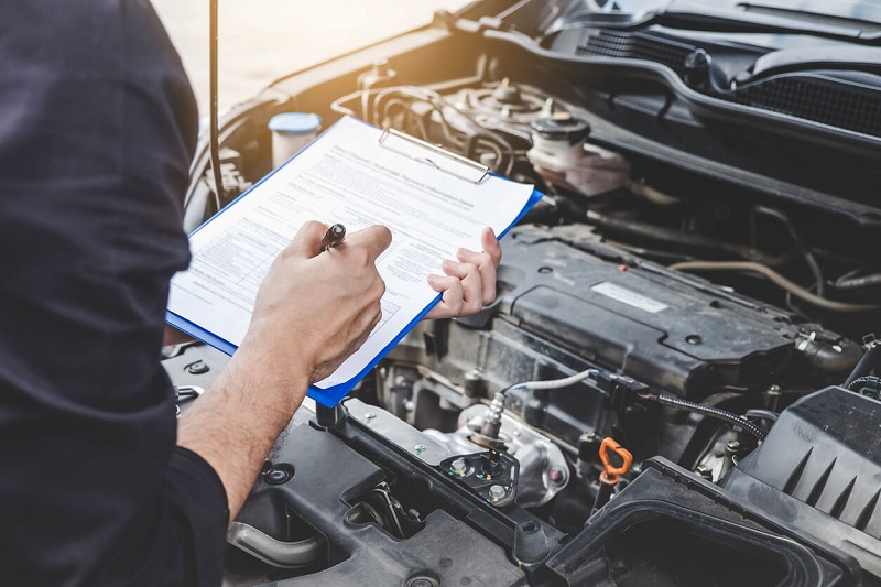 Technician looking at car engine bay holding a clipboard making notes on a roadworthy inspection checklist.