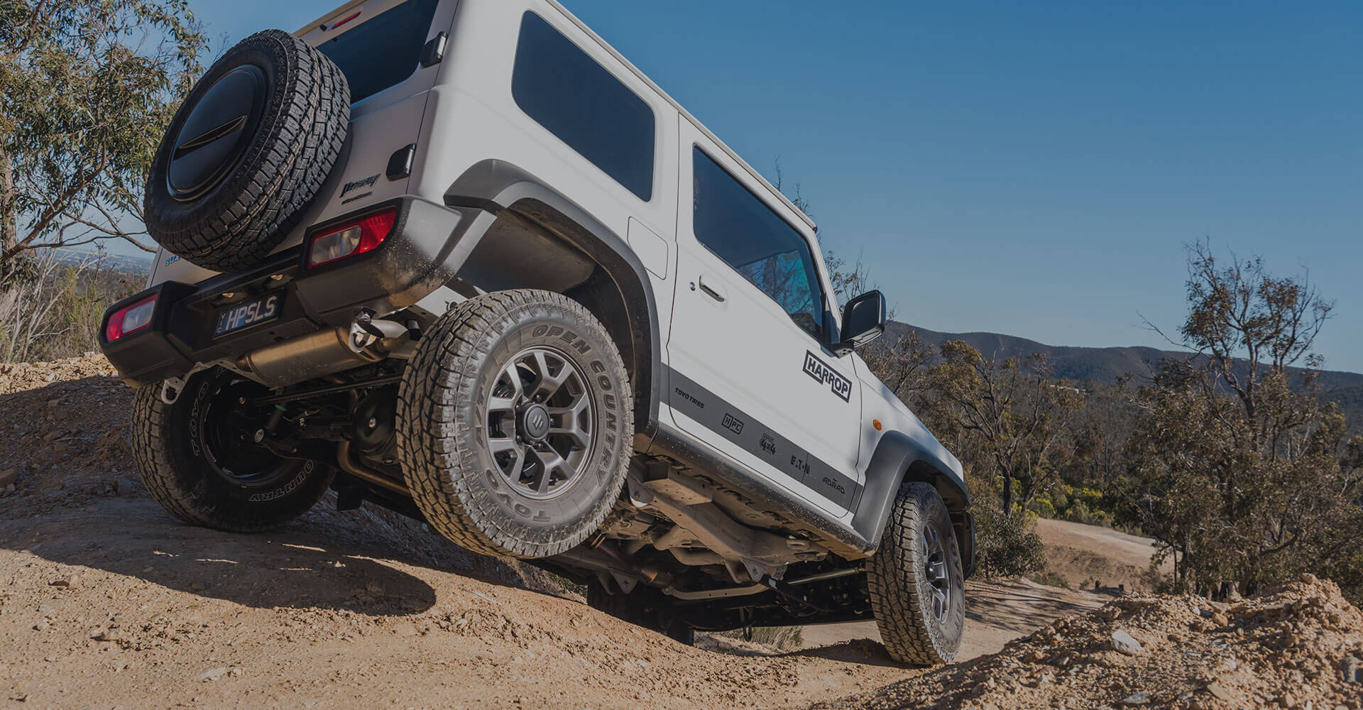 A Suzuki Jimny flexes with the rear wheel off the ground and a set of Toyo Open Country All Terrain tyres on a dusty dirt track.