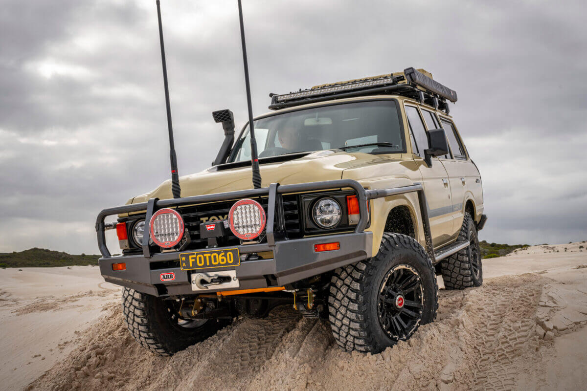 A Toyota 60 series with lots of equipment crawls through the sand and over a dune.