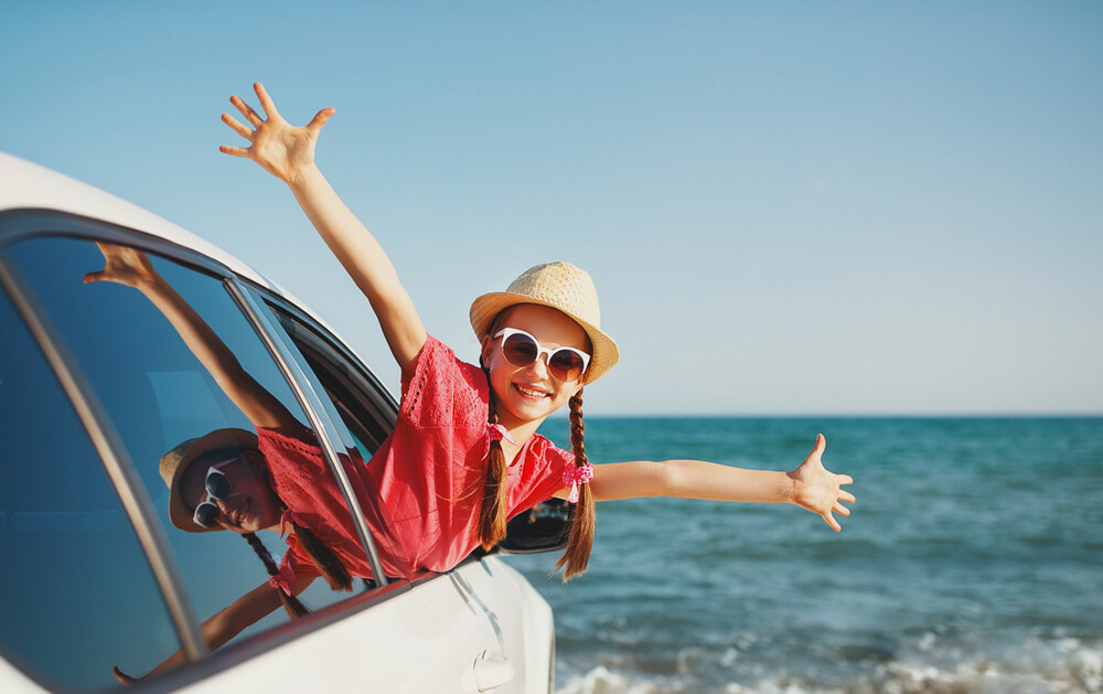 A girl is hanging out the window of a car with her arms wide with a beach in the background.