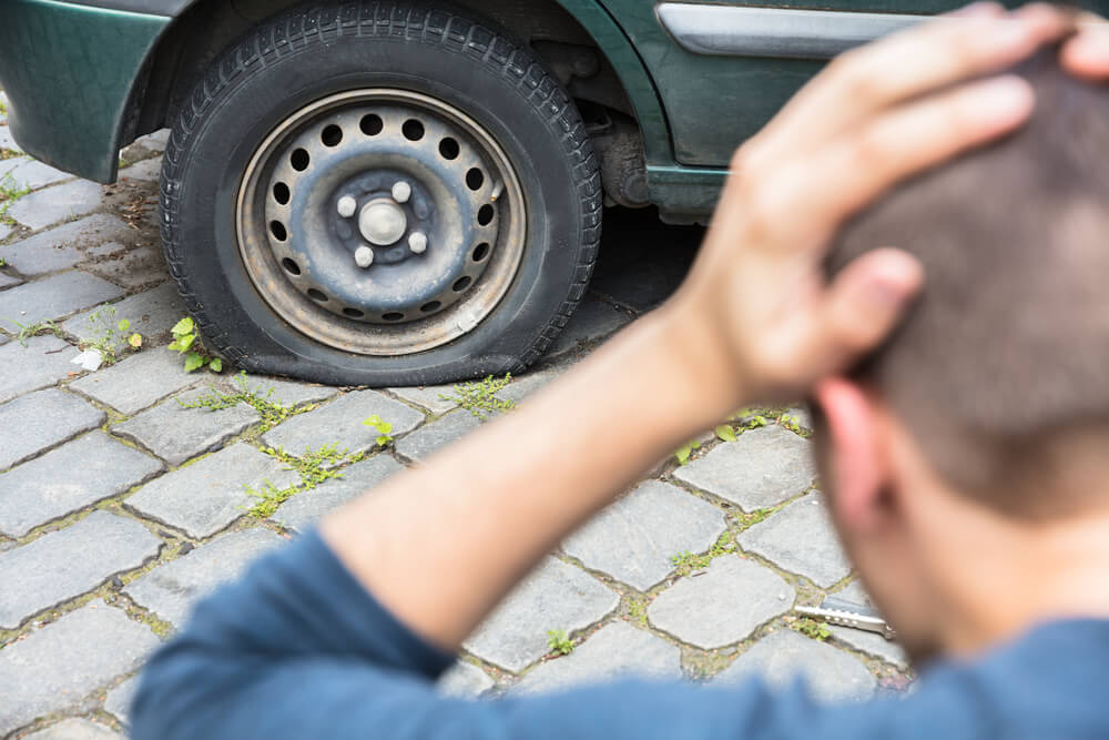 A person with their hands on their head is upset looking at a deflated tyre on their car.