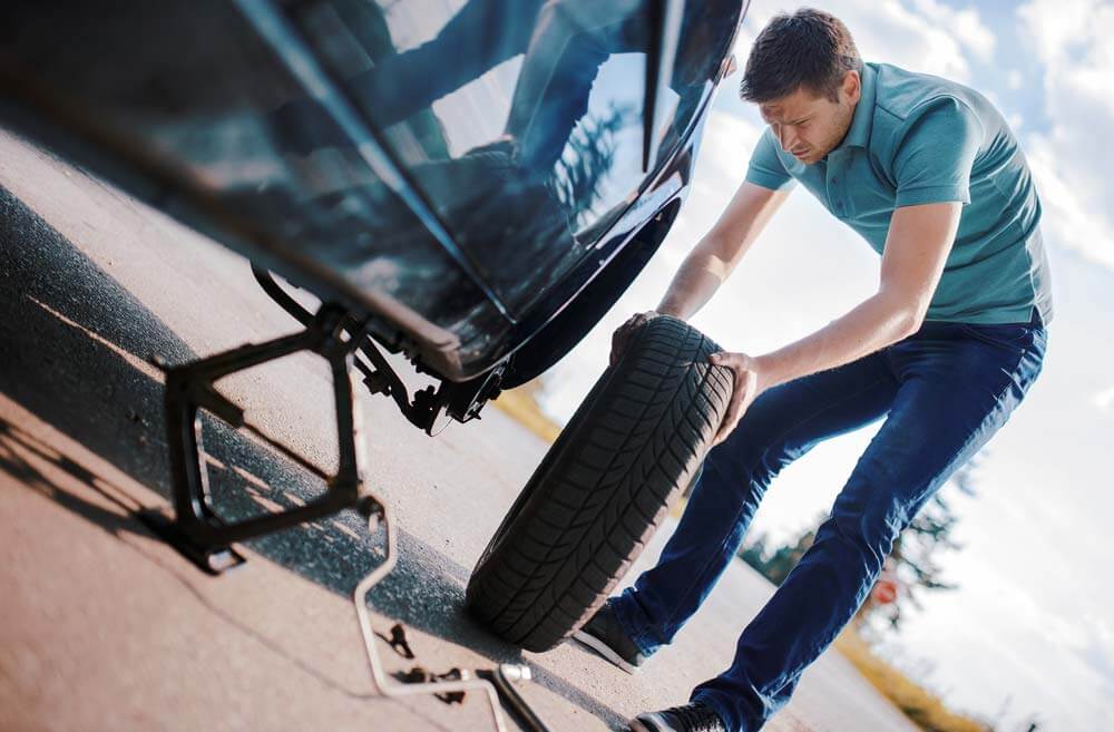 A man pushes the spare tyre up to the car which is lifted by a jack.